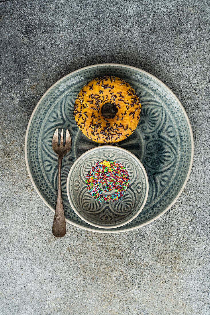 Top view of sweet banana donut placed on gray plate near fork and bowl with colorful sprinkles on gray table