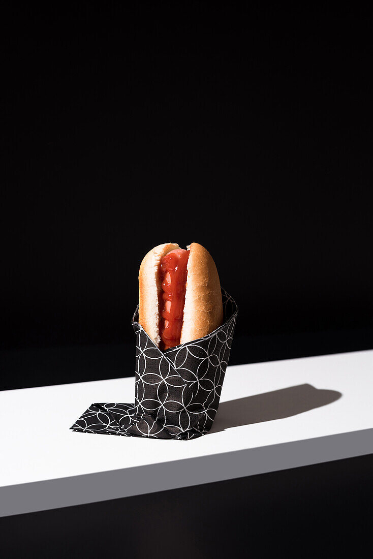 Appetizing bun with sausage and ketchup served table mat over white wooden board against black background in studio