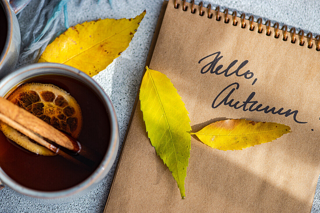 Top view of cup spiced tea with cinnamon sticks and dried orange slices accompanied by bright yellow autumn leaves set beside a notebook with the handwritten words "Hello, Autumn"