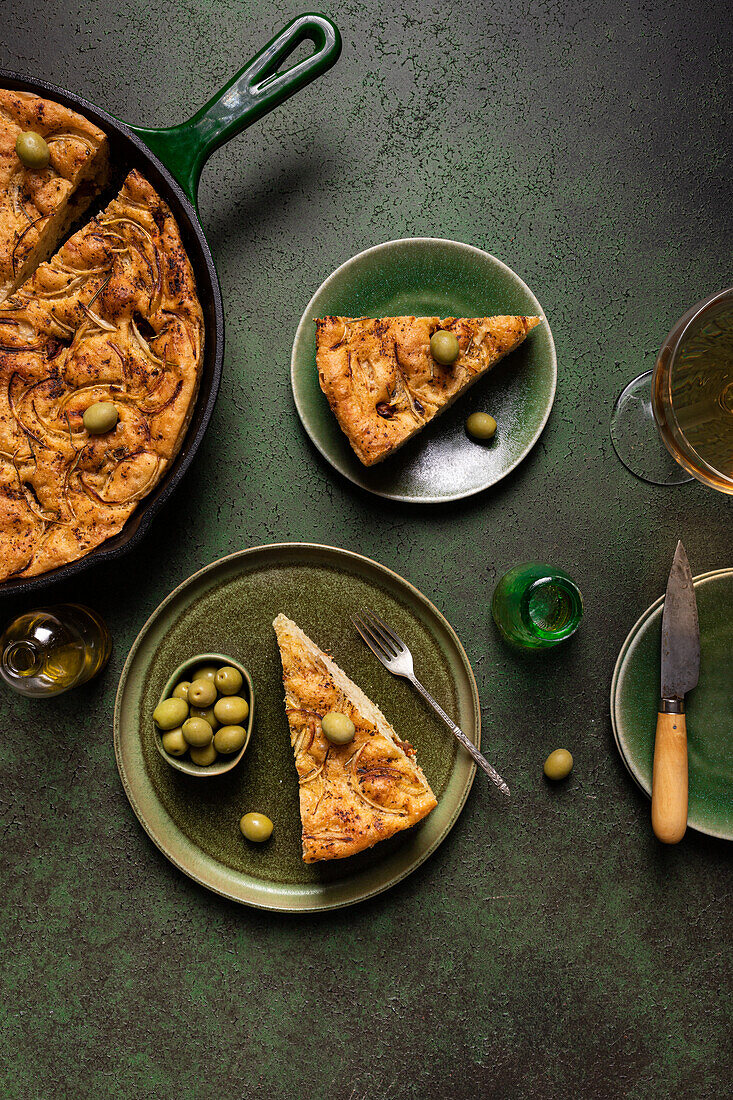 Draufsicht auf eine Scheibe gebackenes italienisches Focaccia-Brot mit grünen Oliven in einem Teller auf einem Esstisch mit Getränk auf grünem Hintergrund