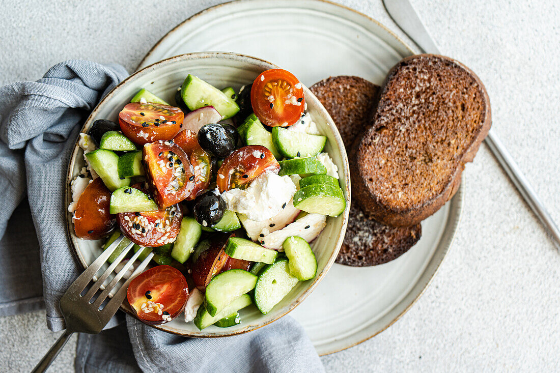 Draufsicht auf eine Keramikschale mit leckerem, gesundem Salat, Gemüse und Brot mit Gurken- und Tomatenscheiben, Besteck und Stoff auf einem grauen Tisch