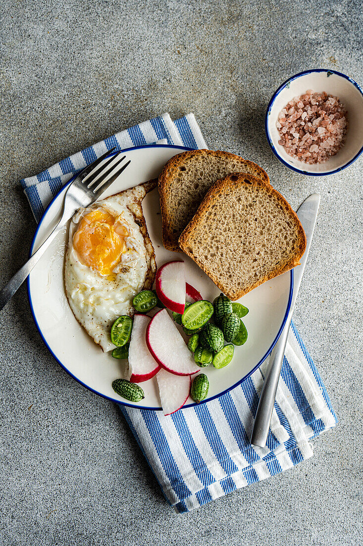 Draufsicht auf eine gesunde Mittagsschüssel mit Brotscheiben, Spiegelei, frischer Gurke, Rettich und Tomate auf einem blau-weiß gestreiften Tuch, daneben eine kleine Schale mit rosa Salz, vor einem grau strukturierten Hintergrund