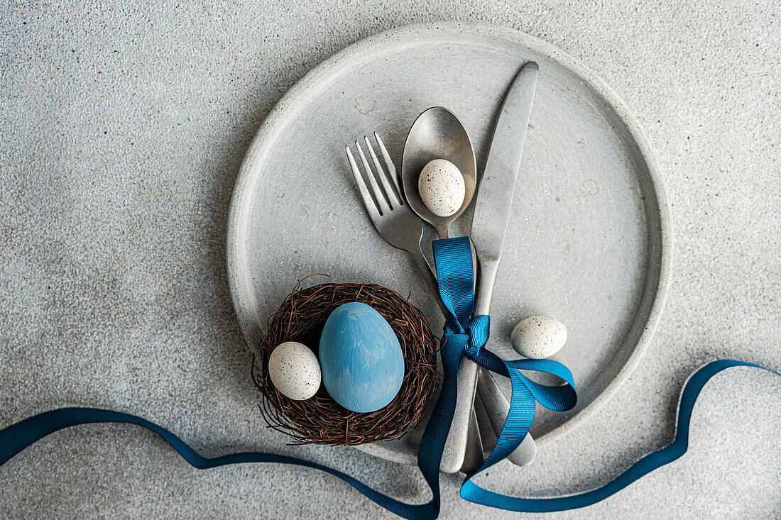 Top view of minimalist yet stylish Easter table setting, with cutlery tied together with blue ribbon on concrete plate, with a small nest with decorative Easter eggs, placed on gray table