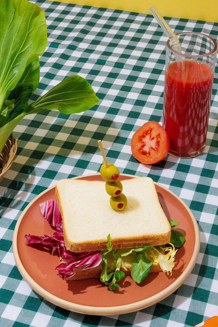 Appetizing healthy sandwich with fresh salad served on plate with olives near glass of tomato juice with glass straw on checkered tablecloth