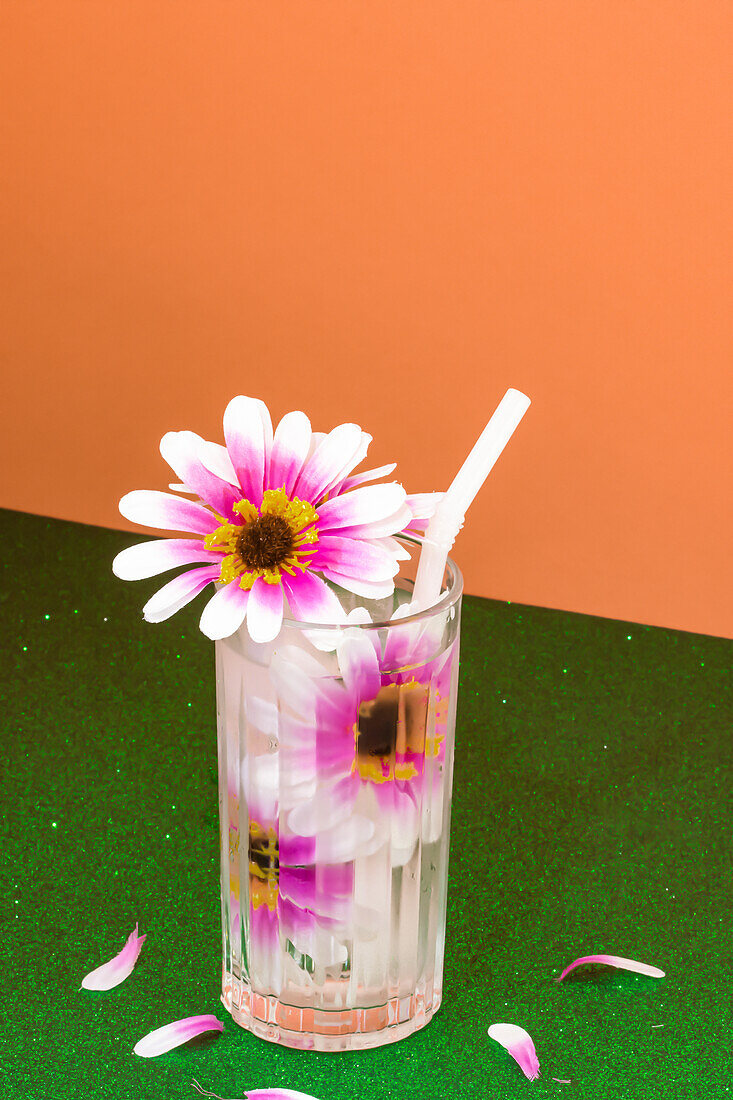 Transparent glass of refreshing cold drink decorated with pink flowers and straw placed on green surface against bright orange wall