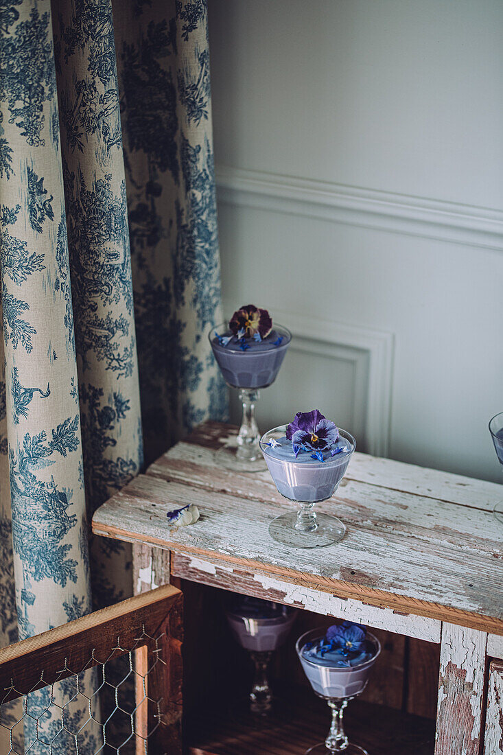 Verschiedene Gläser mit schmackhaftem, blauem Spirulina-Pudding, dekoriert mit Blumen, auf einem schäbigen Holzschrank