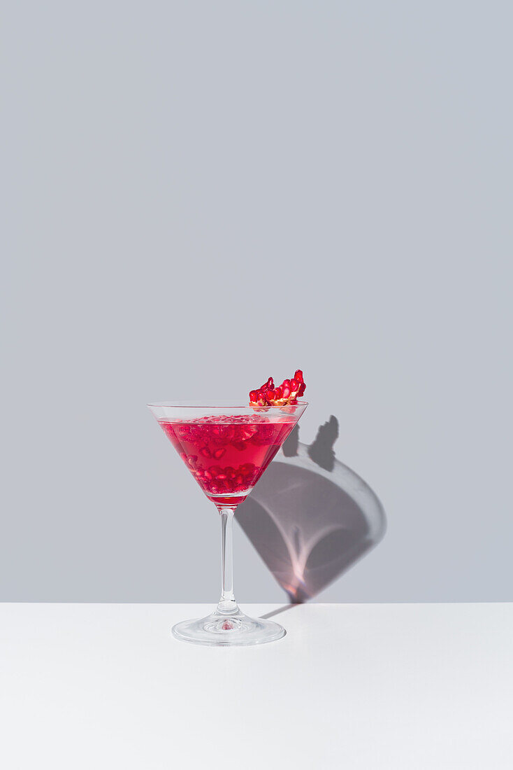 Glass filled with red pomegranate cocktail served with pomegranate seeds against a muted gray backdrop, casting a soft shadow