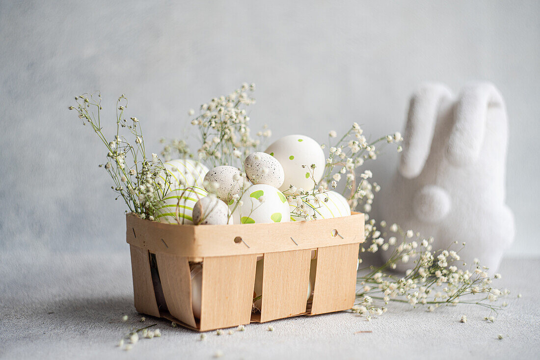 A selection of decorated Easter eggs nestled in a wooden basket alongside sprigs of gypsophila, with a soft gray background