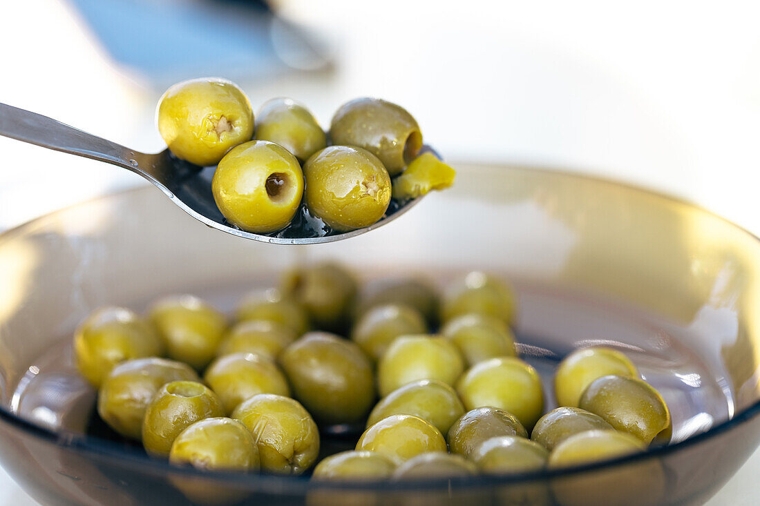 A spoonful of pitted green olives is held above a glass bowl filled with a similar assortment against a neutral background
