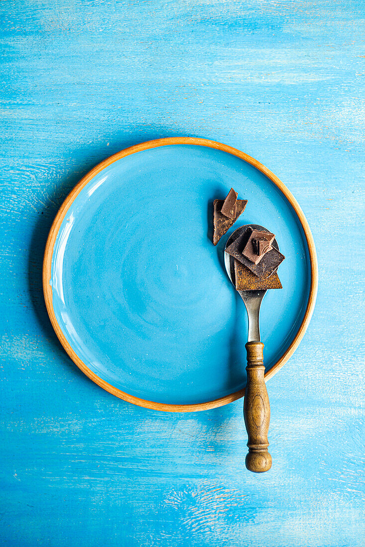 Draufsicht auf Stücke verschiedener Schokoladensorten auf einem Löffel und einem blauen Keramikteller vor blauer Oberfläche