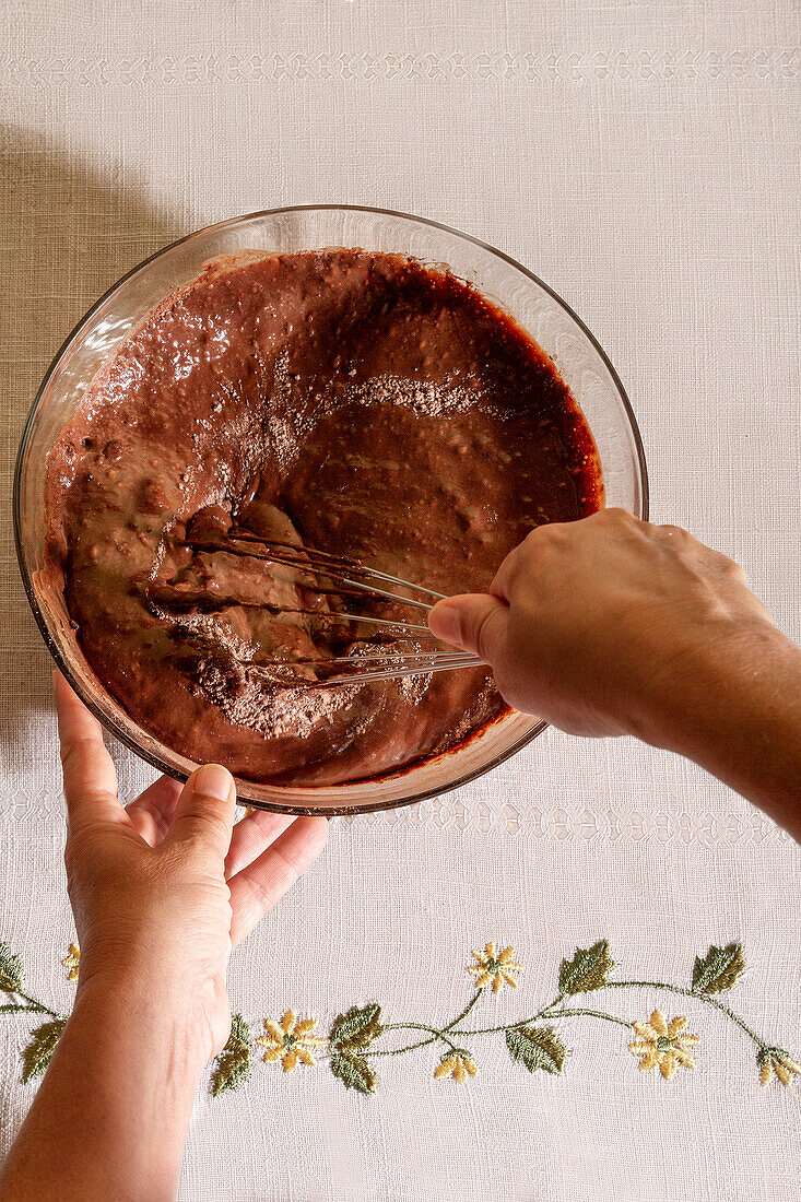 Top view of Hands of anonymous person whisking chocolate batter in a clear glass bowl on a floral embroidered cloth