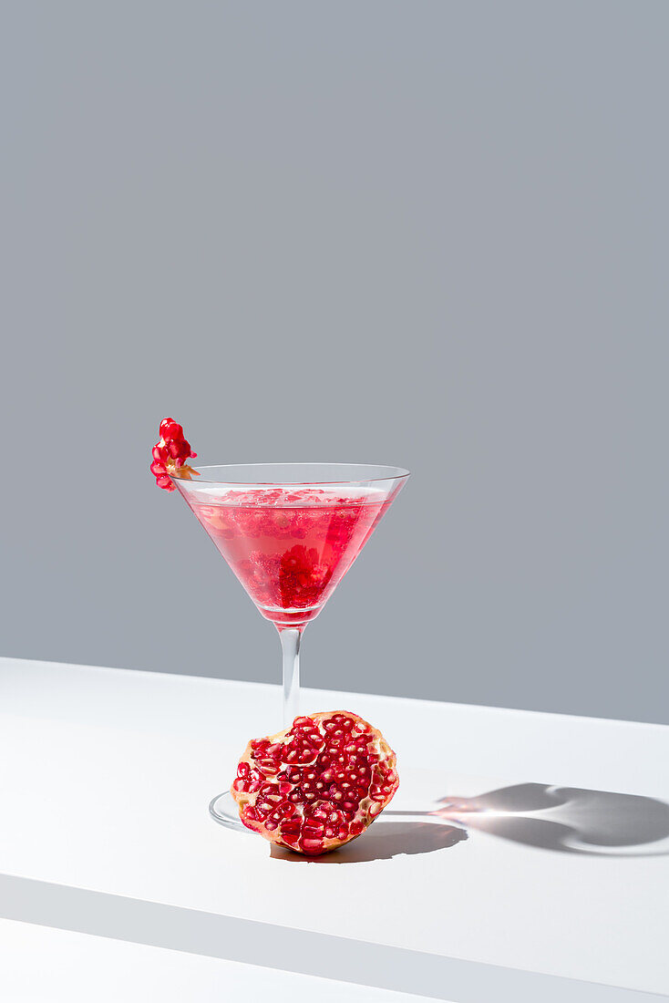 Glass filled with red pomegranate cocktails served with pomegranate seeds next to a juicy fresh pomegranate, against a gray backdrop, casting a soft shadow