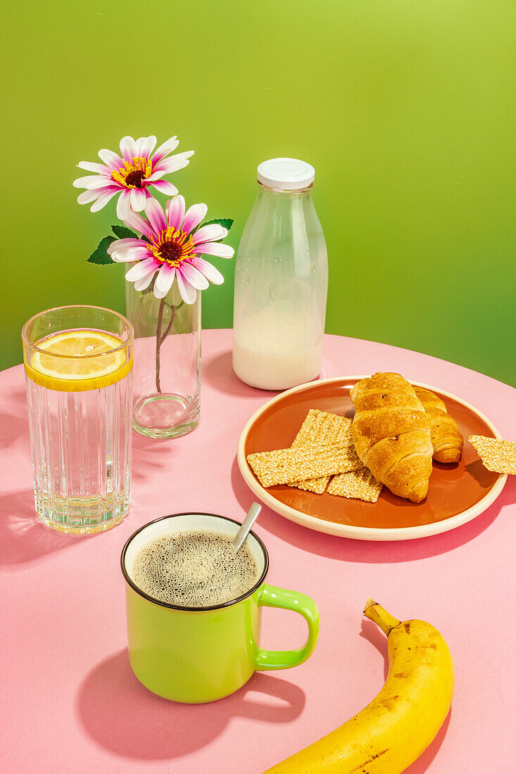 Appetizing croissants and banana placed near glass bottle of milk and water with lemon slice against flowers in vase and cup of aromatic coffee on pink table against green background