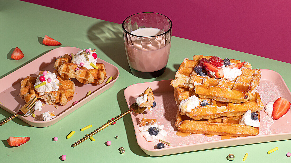 From above tasty sweet waffles topped with berries fruits sauce and cream served on pink tray on colorful table background near glass with hot milk beverage in light kitchen