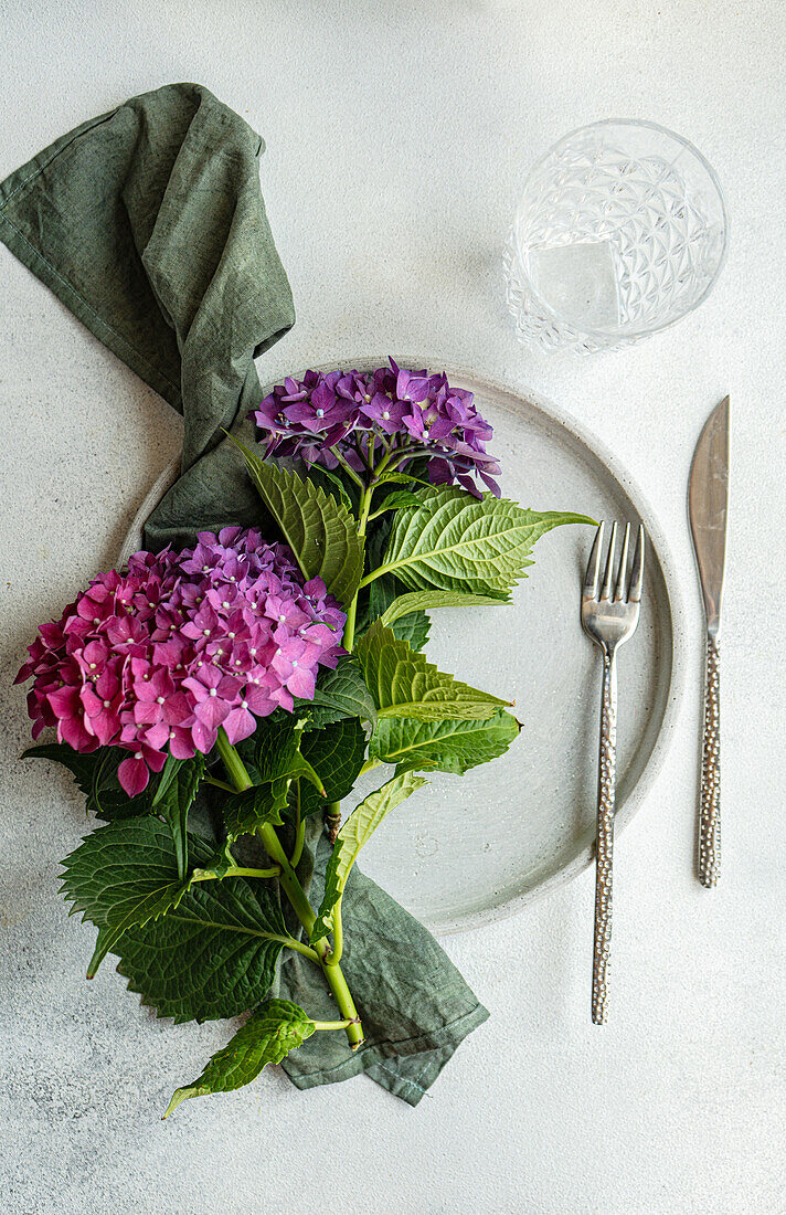 Draufsicht auf eine violette Hortensie, die auf einem weißen Tisch neben Keramiktellern und Gläsern steht