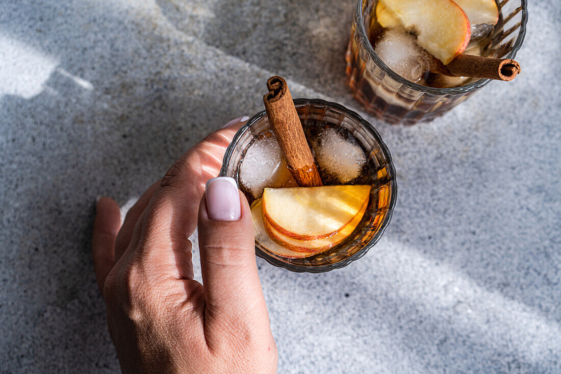 From above of anonymous hand holding a glass of apple cider cocktails adorned with cinnamon stick, star anise, and apple slices, showing fresh ice cubes