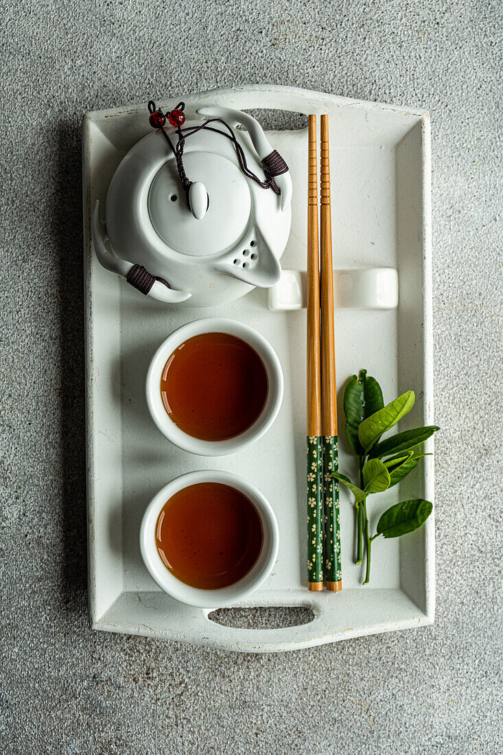 Top view of tea set in Asian style with lemon leaves and chopsticks placed on white tray against gray background