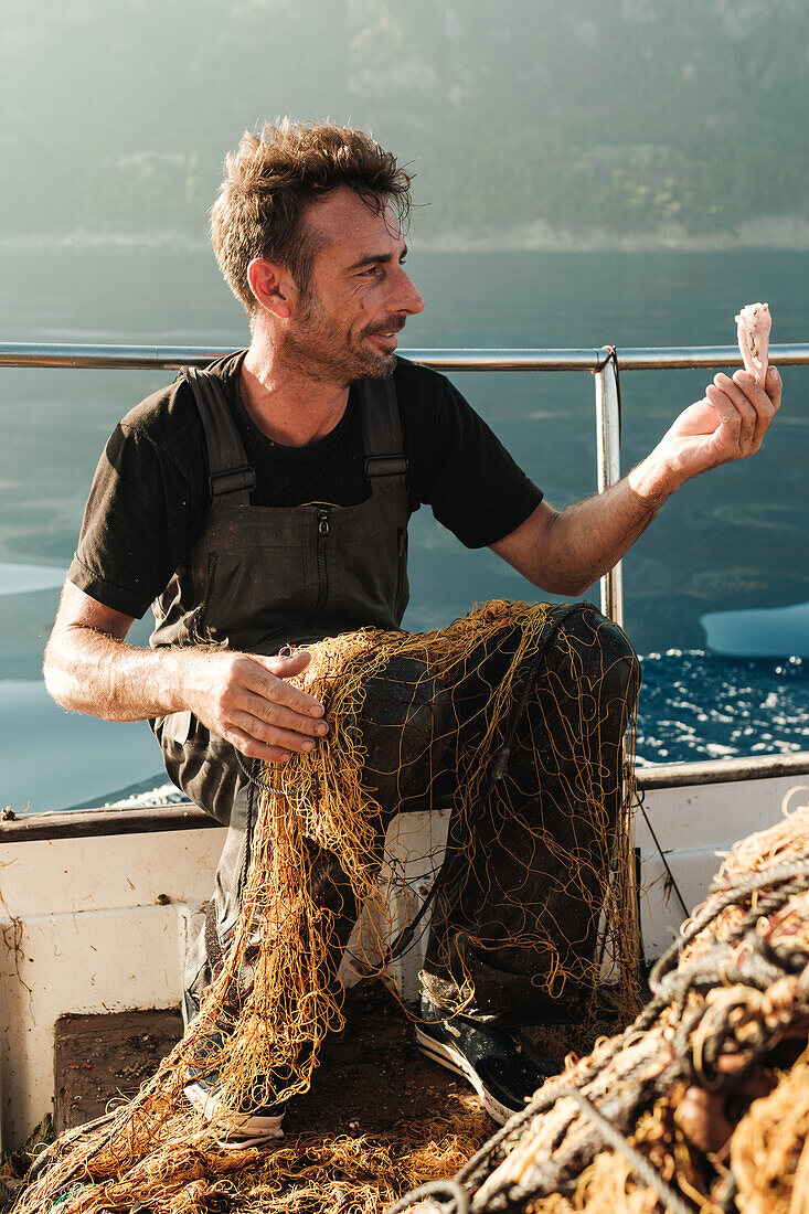 Ganzkörperaufnahme eines lächelnden männlichen Fischers, der beim traditionellen Fischfang Muscheln aus dem Netz fischt, vor einer Meereslandschaft in Soller auf der Baleareninsel Mallorca