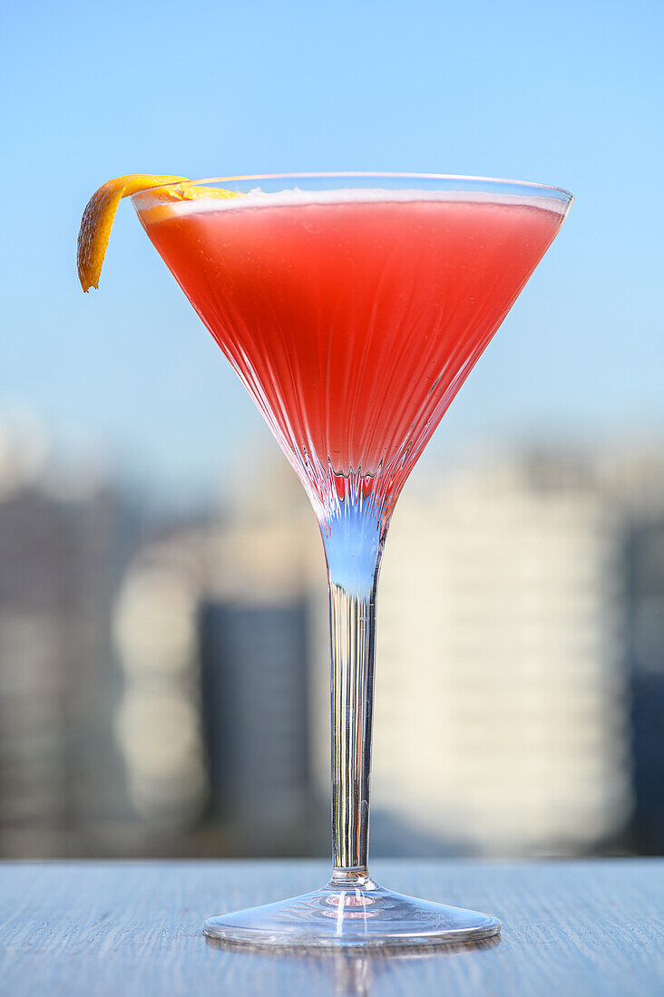 Martini glass filled with red alcoholic cosmopolitan cocktail garnished with lemon peel placed against blurred cityscape