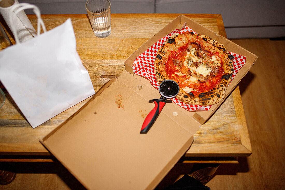 Blick von oben auf eine appetitliche gebackene Pizza mit Tomaten und Käse mit knuspriger Kruste, die in einem Karton mit Messer auf dem Tisch liegt