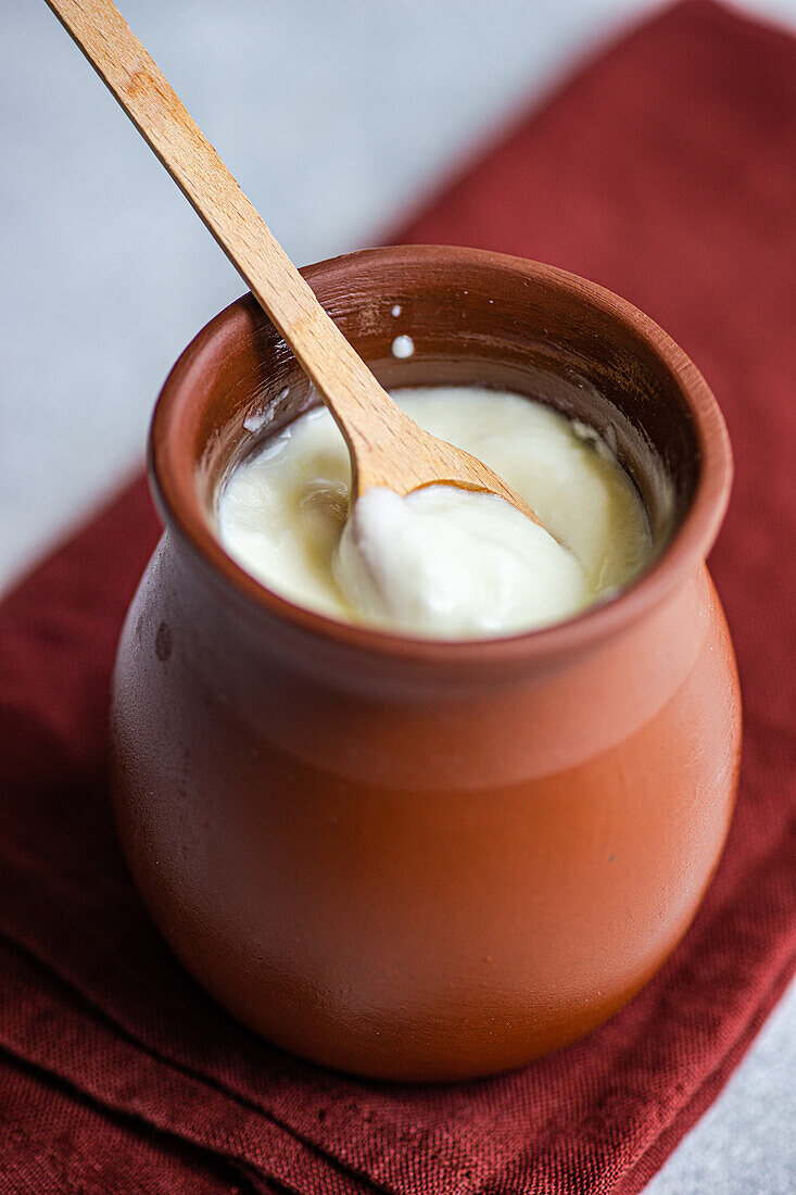 Traditioneller saurer georgischer Joghurt, bekannt als Matsoni, in einem Tontopf mit Holzlöffel auf einer braunen Serviette vor einem unscharfen Hintergrund, von oben betrachtet