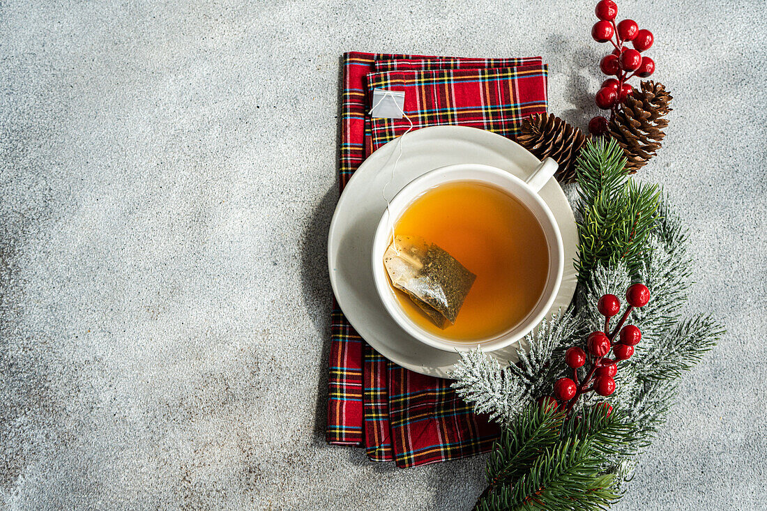 Draufsicht auf eine weihnachtliche Tasse Tee auf einer roten Serviette neben Stechpalmen und Tannenzapfen auf einem grauen Tisch