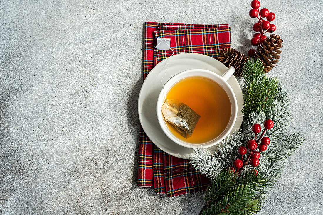 Top view of Christmas cup of tea placed on red napkin near holly and pine cones on gray table