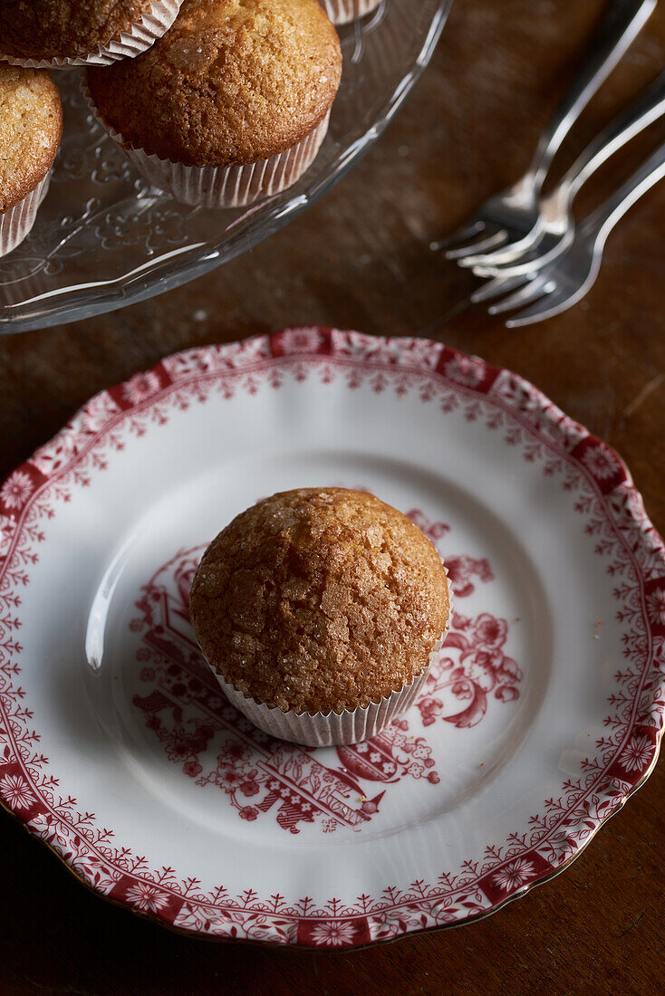 A single golden-brown muffin, sprinkled with sugar sits atop a red and white vintage-patterned plate, with a cake stand and forks in the background
