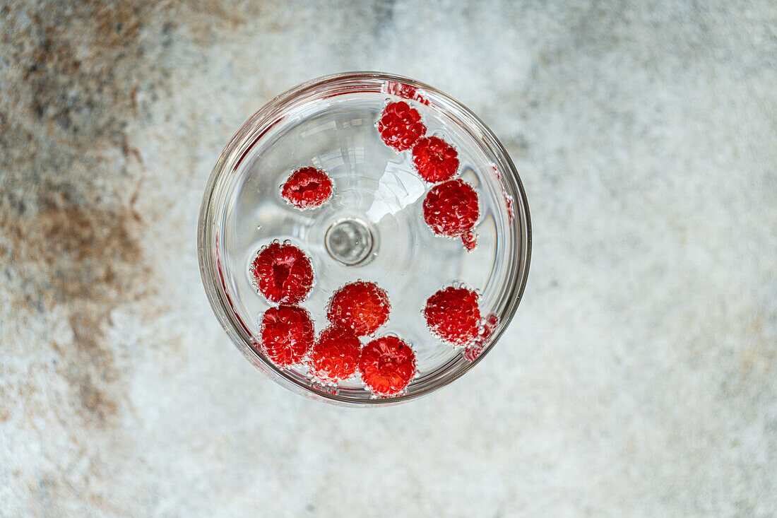 Draufsicht auf einen transparenten, glatten Glasbecher, gefüllt mit erfrischendem Cocktailsekt mit reifen frischen Himbeeren vor unscharfem Hintergrund