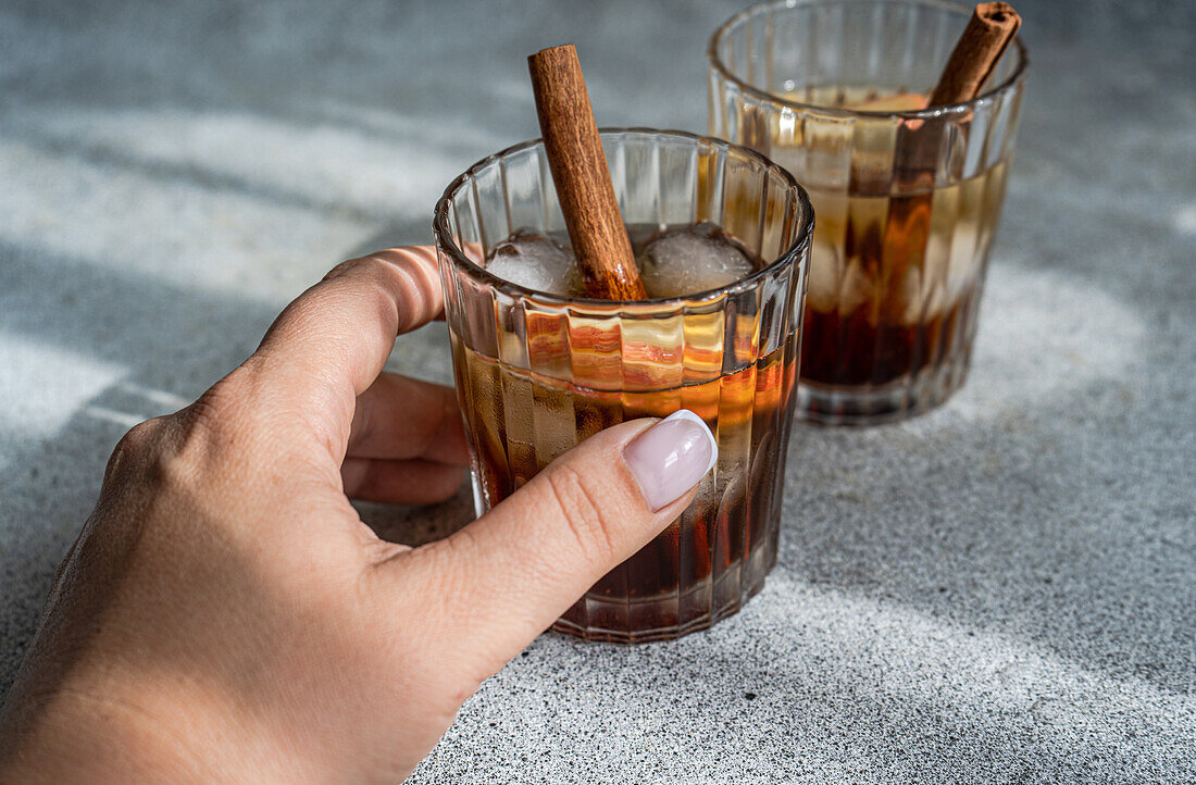 Anonymous hand holding a glass of apple cider cocktail adorned with cinnamon stick, star anise, and apple slices, showing fresh ice cubes