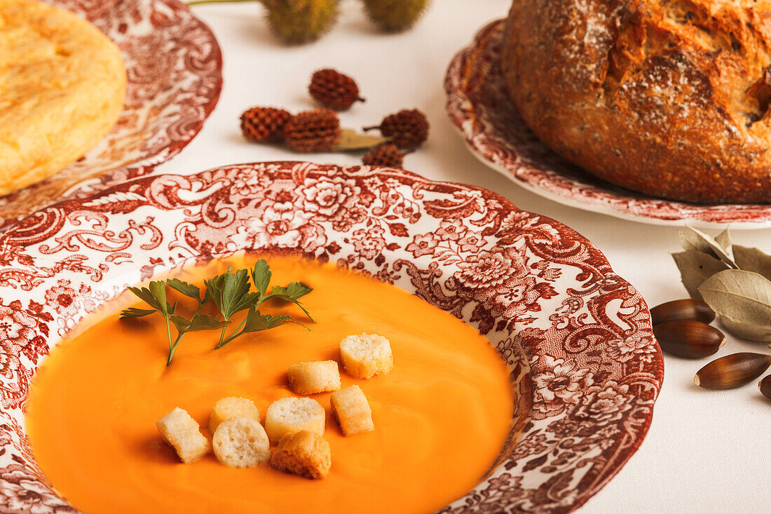 A heartwarming serving of salmorejo garnished with croutons and parsley, accompanied by a rustic loaf of bread, all laid out on decorative plates.