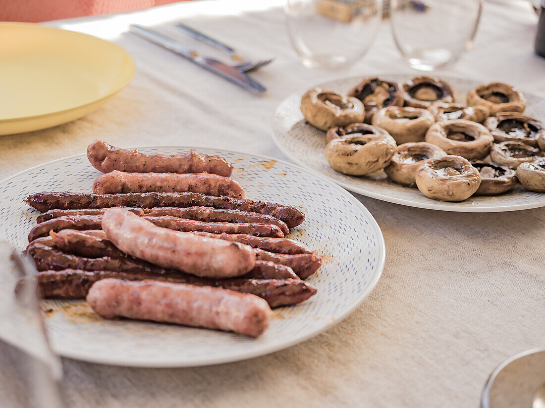 Hoher Blickwinkel auf köstliche gegrillte Hot Dogs und weiße gefüllte Pilze auf einem Teller mit Glasschalen über einer Tischdecke auf einem Esstisch bei Tageslicht