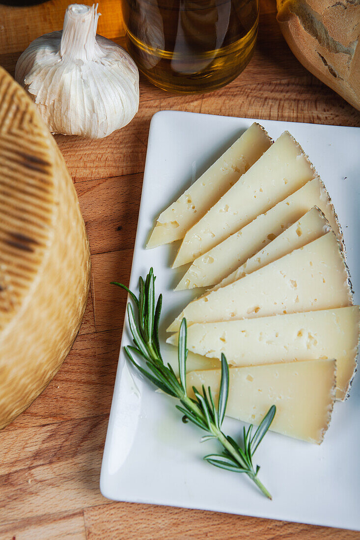 Handwerklich hergestellter Käse in Scheiben auf einem weißen Teller, akzentuiert mit einem Zweig frischen Rosmarins, mit Knoblauch und Olivenöl im Hintergrund