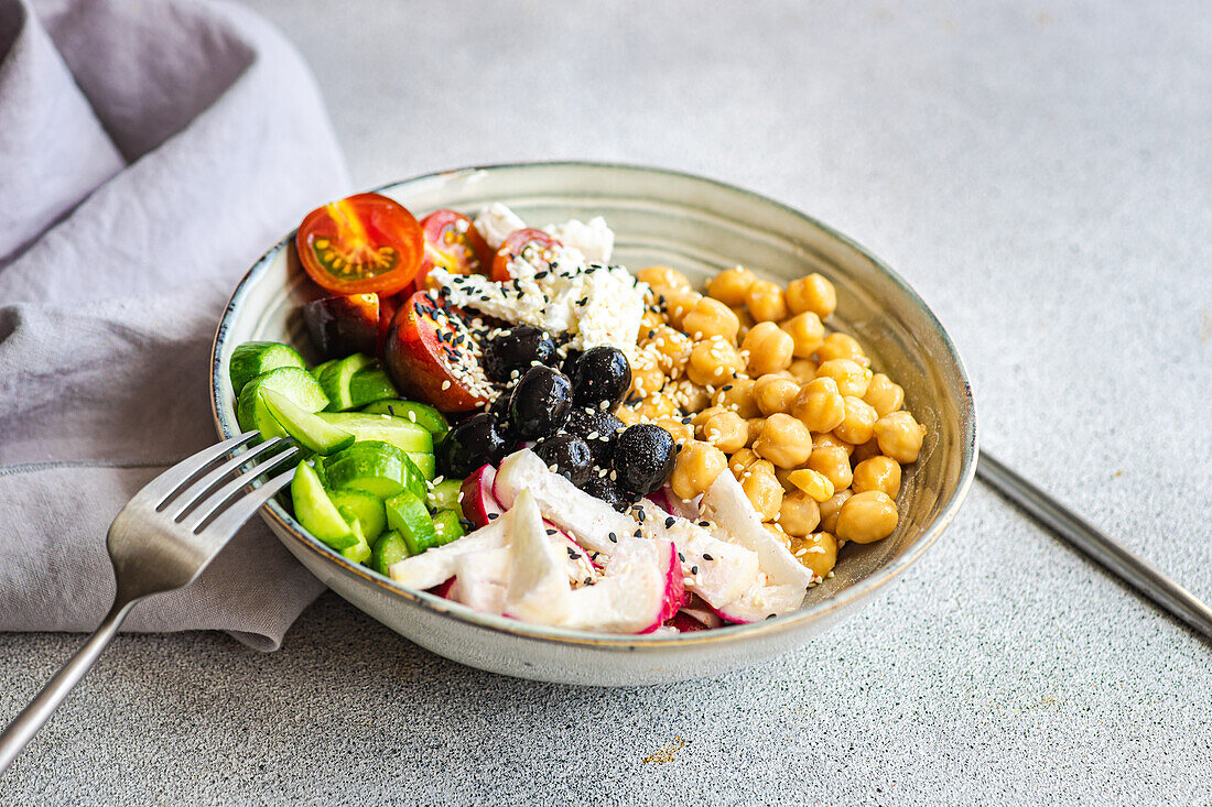 High angle of bowl with healthy salad and vegetables with chickpea sesame seeds and olives with cutlery and fabric placed on bright gray table
