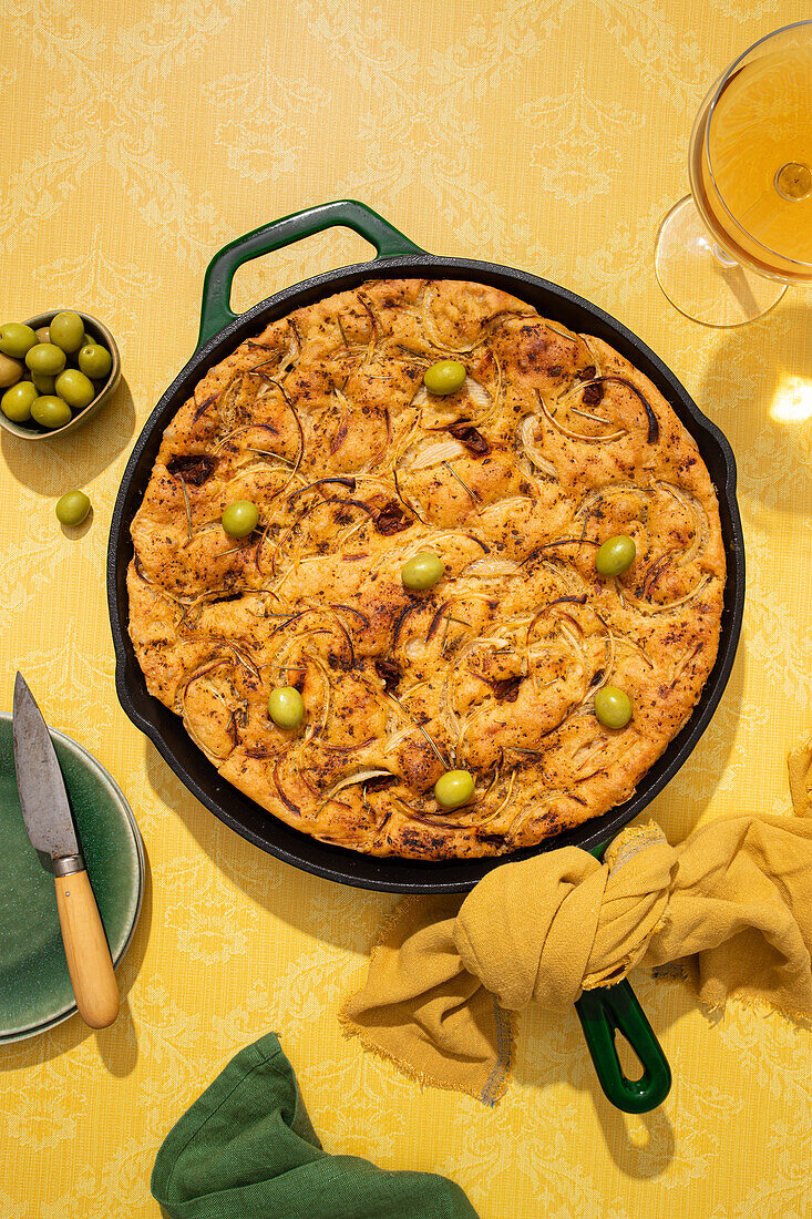 Draufsicht auf flaches, gesäuertes, im Ofen gebackenes italienisches Focaccia-Brot in einer Pfanne, serviert mit frischen grünen Oliven und Honig in Schalen auf gelber Hintergrundfläche