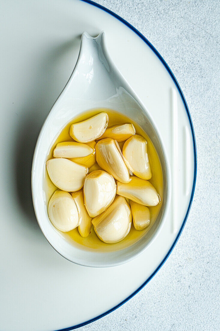 Top view of peeled and baked garlic cloves submerged in oil, showcased in a ceramic spoon placed on a white plate with a subtle blue border, against a light gray background