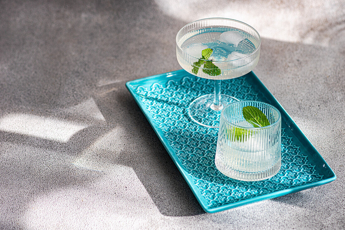 Zwei stilvolle Cocktailgläser auf einem blauen, strukturierten Serviertablett, verziert mit Minzblättern, vor einem weichen, schattenhaften Hintergrund