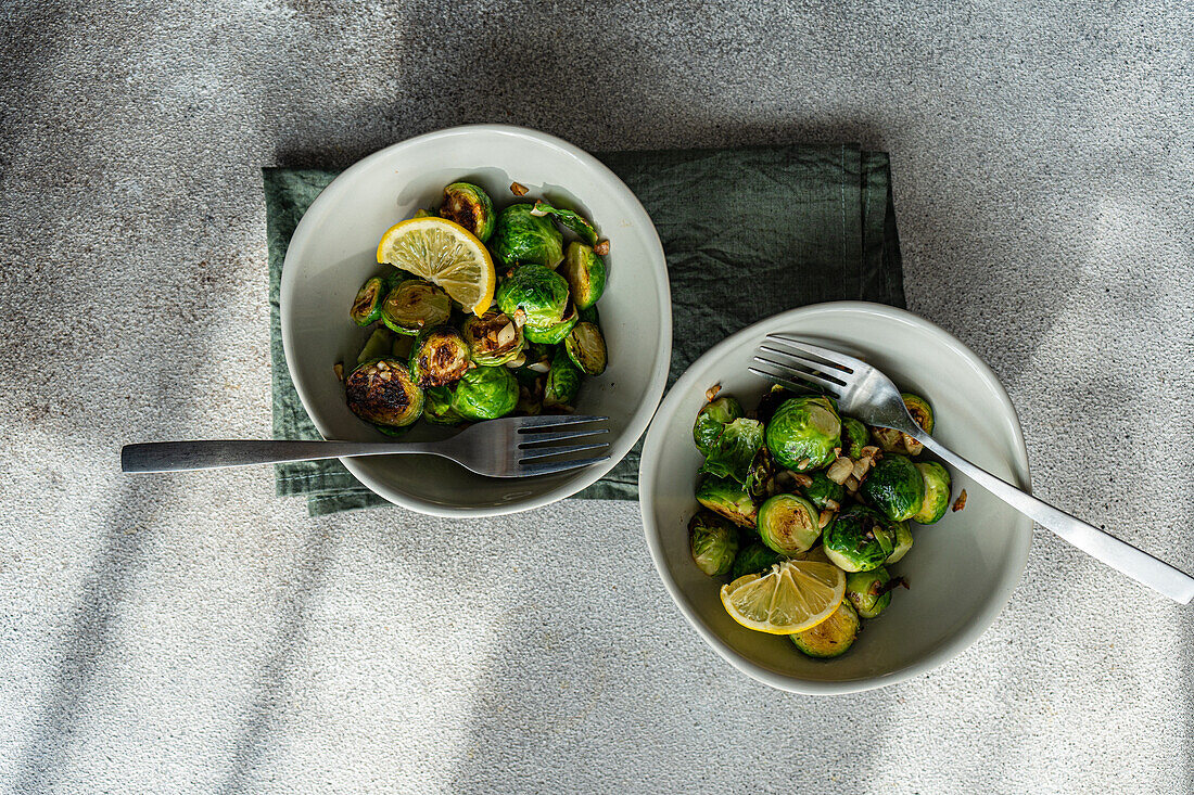 Top view of two bowls filled with barbecued Brussels sprouts flavored with garlic and spices, accompanied by silver forks on a textured tablecloth