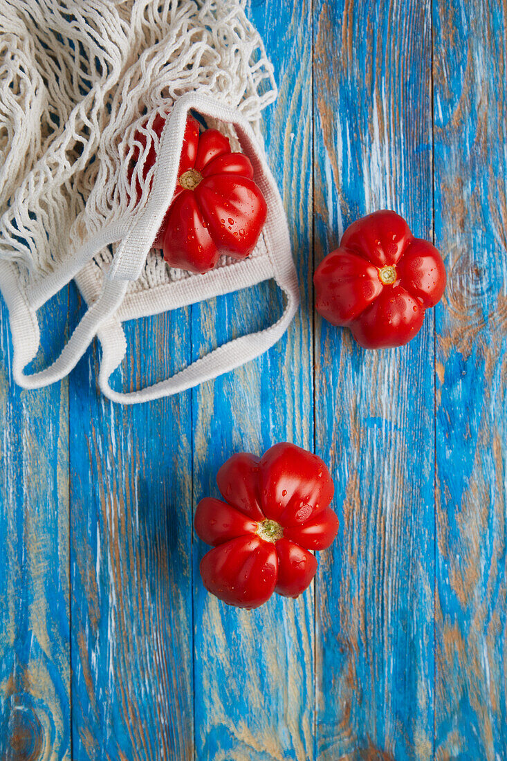 Draufsicht auf frische reife rote Tomaten mit umweltfreundlichem Baumwollnetzbeutel auf blauem Holztisch