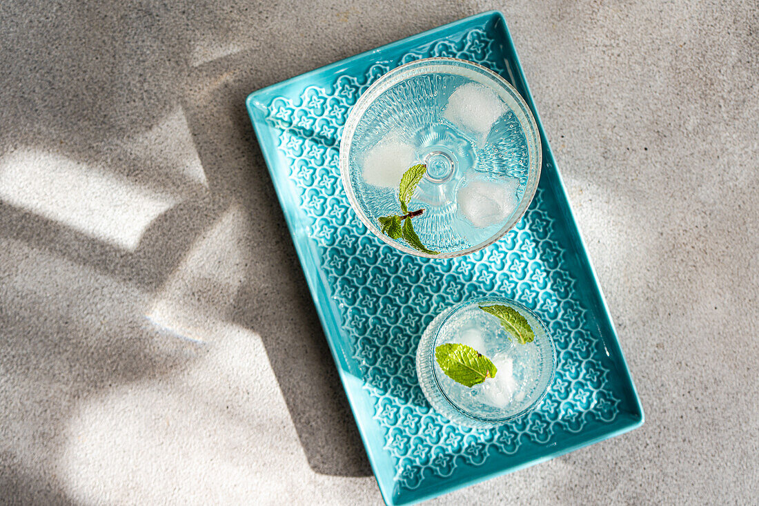 Two gin and tonic cocktails with lime and ice on a decorative blue tray, casting shadows in natural light