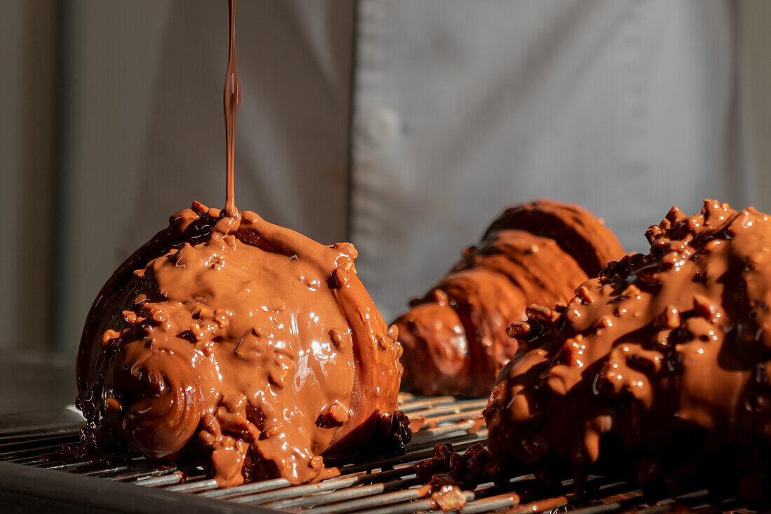 Unbekannte Person gießt Schokoladenglasur auf appetitliche Croissants, die auf einem Metalltablett liegen, während sie in einer Fabrik Gebäck zubereiten