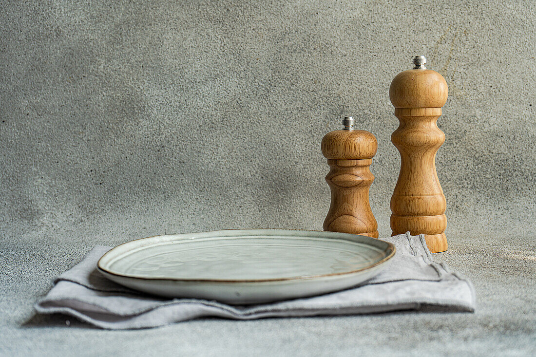 Ansicht von oben auf einen Keramikteller mit Löffel und Serviette neben einer Gabel auf einer grauen Fläche am Küchentisch für eine Mahlzeit auf einem Betonhintergrund