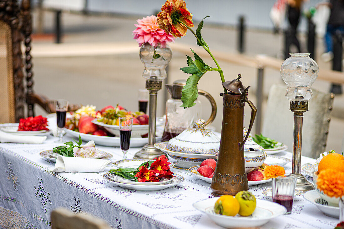 An elegant outdoor dining table adorned with fresh fruits, ornate dishes, vintage lanterns, a brass pitcher, and a vibrant dahlia flower on a lace tablecloth.