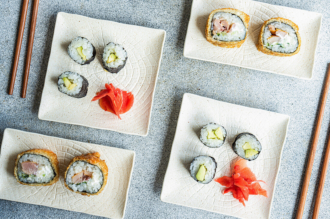 Verschiedene Sushi-Rollen auf Tellern mit Stäbchen und Sojasauce auf weißem Hintergrund von oben