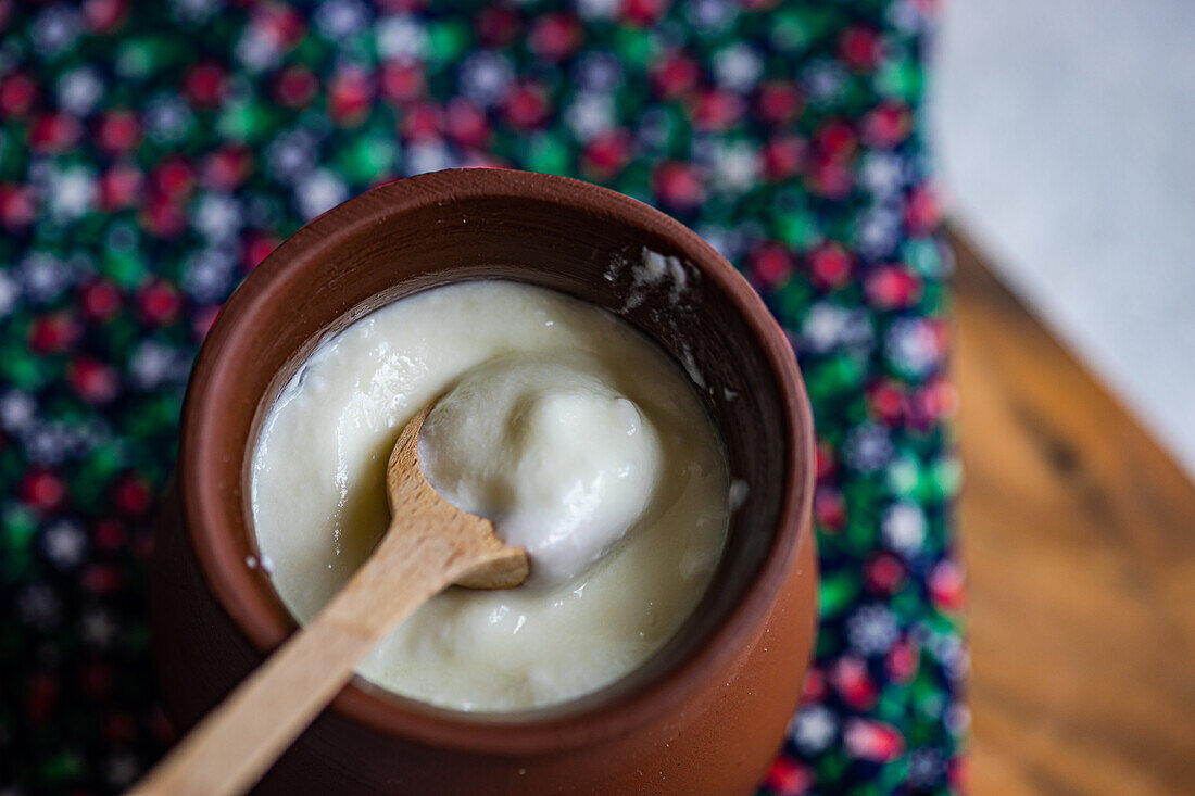 High angle of traditional serving of Georgian sour yogurt known as Matsoni in clay pot with wooden spoon on colorful napkin against blurred background