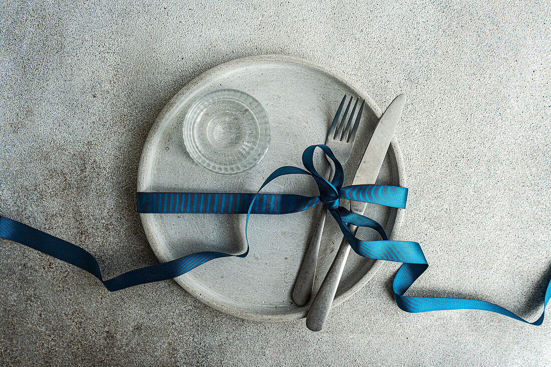 Draufsicht auf einen minimalistisch und doch stilvoll gedeckten Ostertisch mit einem grauen Teller, der mit einem blauen Band gebunden ist, auf einer grauen Fläche