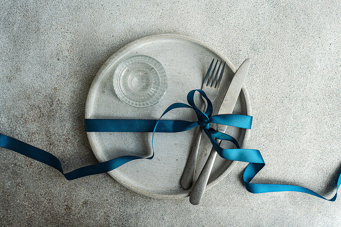 Top view of minimalist yet stylish Easter table setting, with a concrete grey plate bound by a rich blue ribbon, grey cutlery is neatly tied together, while a textured clear glass sits at the center