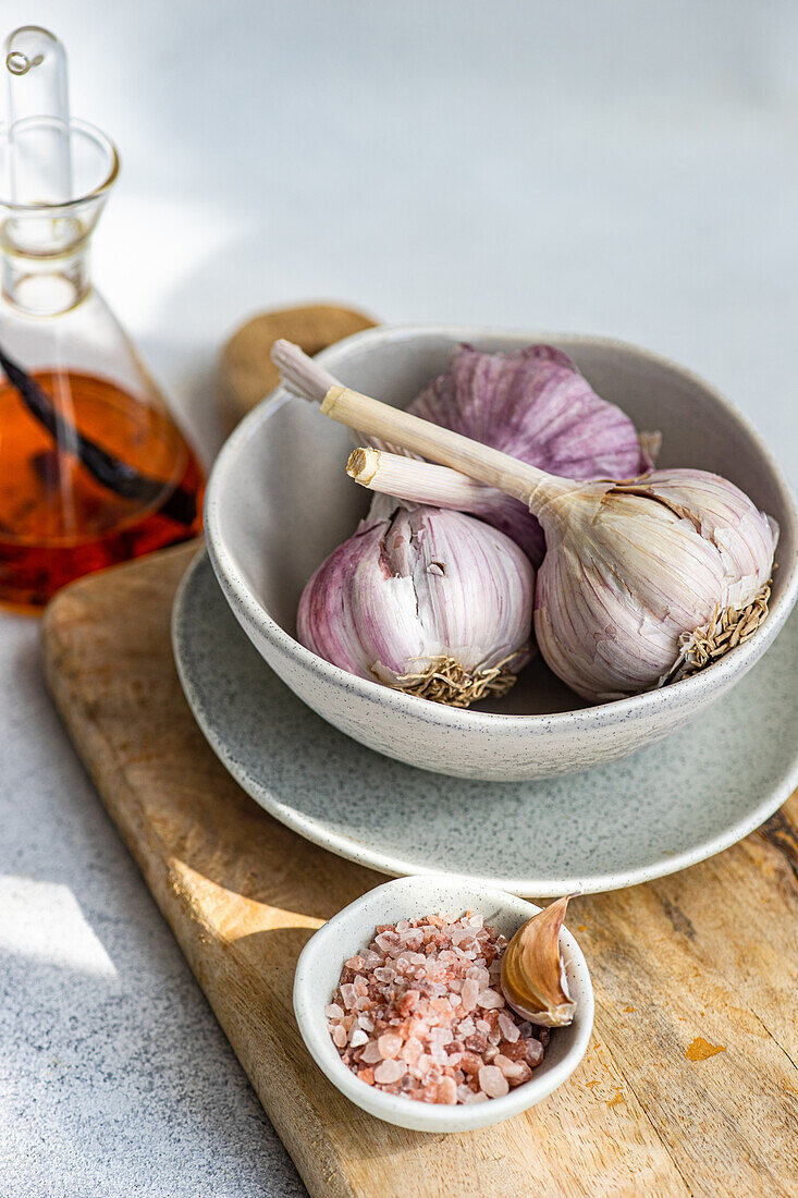 Raw garlic bulbs in a ceramic bowl next to a decanter of amber liquid set on a wooden board with a dish of pink salt and a garlic clove