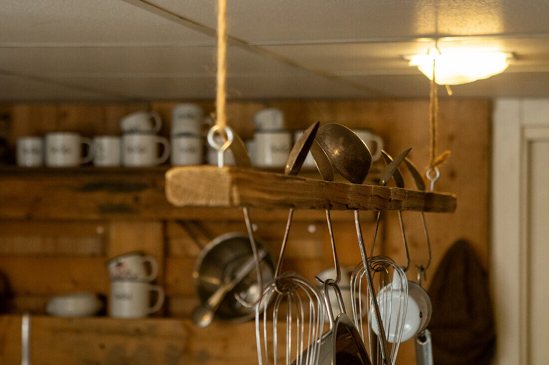 Variety of kitchen utensils hanging from a wooden shelf, with an array of white soup mugs in the background, softly illuminated by overhead lighting