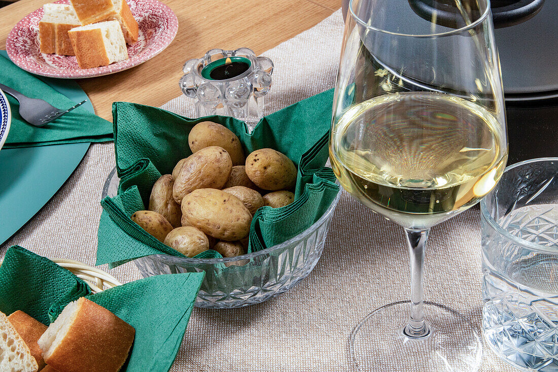 Ein gedeckter Tisch mit einem Glas Weißwein, frischem Brot und einer Schüssel mit gebackenen Kartoffeln, bereit für eine Dinnerparty